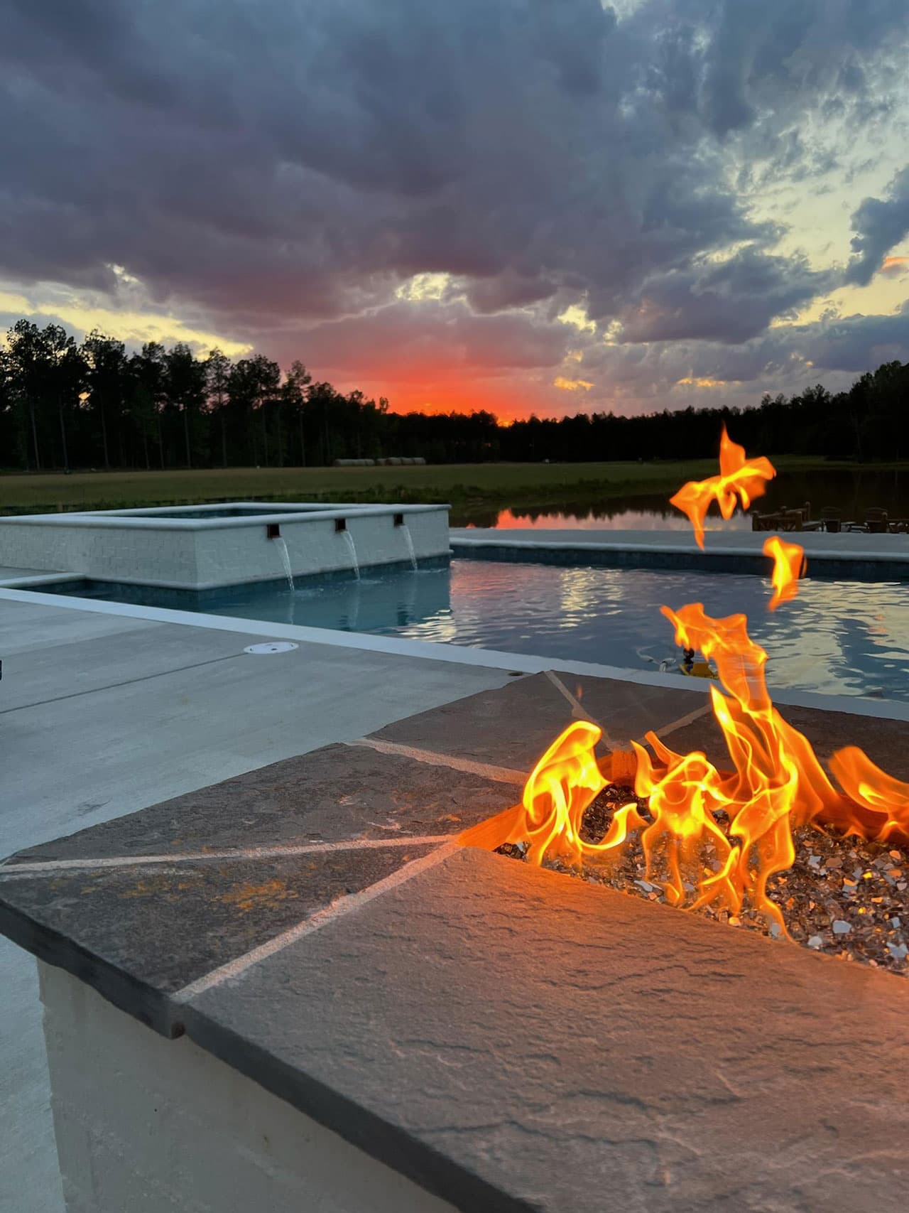 A raised spa spilling into a private pool, with a stone fire pit burning in the foreground and a hay field and pine treeline behind
